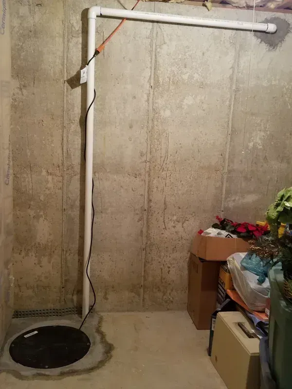 A white sump pump discharge pipe rising from a basement floor cover and extending horizontally through a concrete wall.