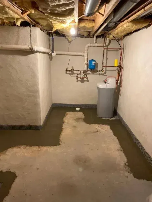 A basement corner with unfinished concrete walls, exposed ceiling joists, utility pipes, and a water softener system.