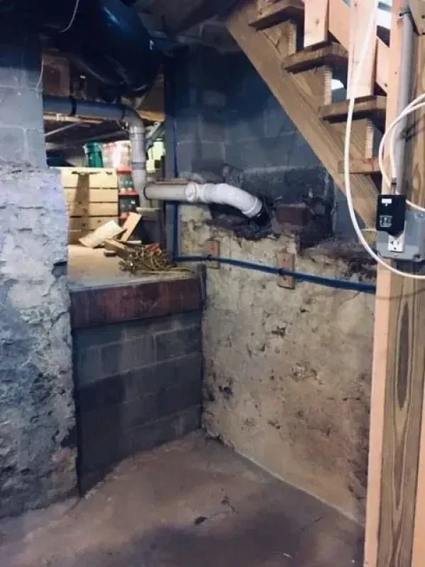 A dark basement corner with stone walls, wooden stairs, white plumbing pipes, and a blue line running along the wall.