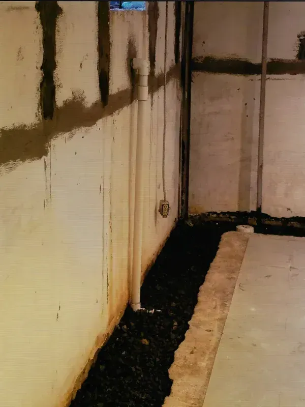 A basement foundation wall showing a vertical white pipe extending into a trench filled with black stone.