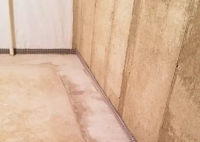 Basement corner showing a concrete wall with insulation panels and a perforated drainage channel along the floor.