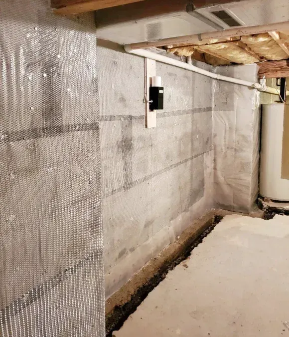 A concrete basement wall partially covered with plastic vapor barrier sheeting, next to a white plastic drainage channel.