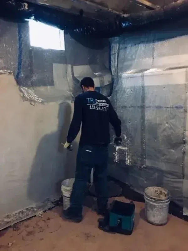 A worker in a dark shirt applies sealant to a concrete wall in a basement next to two buckets and plastic sheeting.
