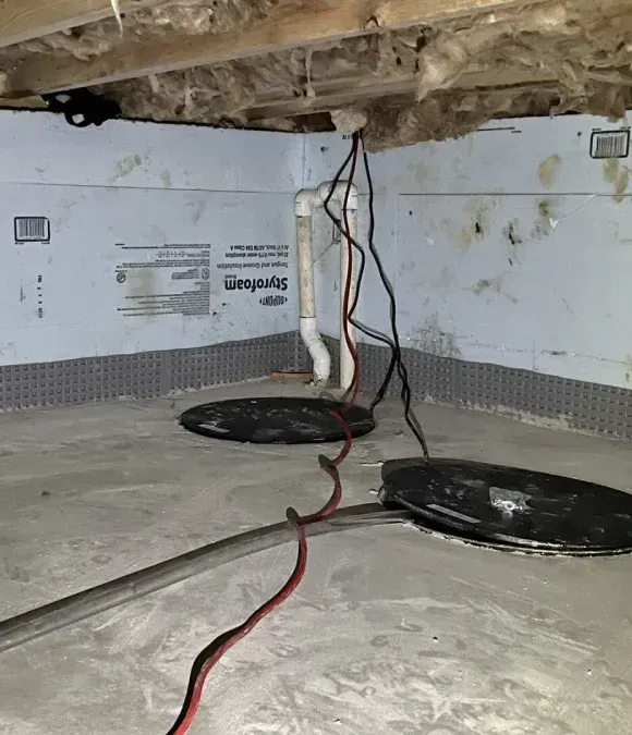 Two sump pump basins set in a crawlspace with insulation on the walls and electrical cords running up to the floor joists.