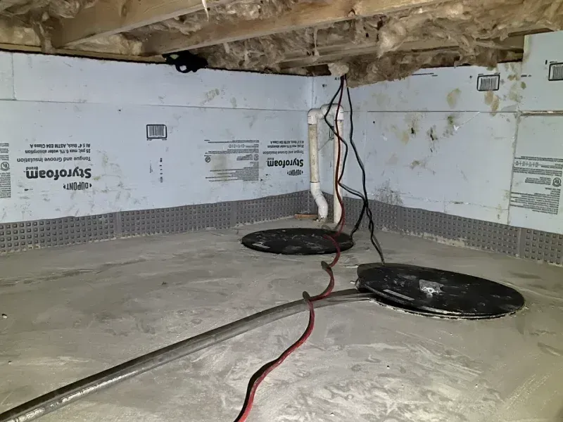 Crawl space with two covered sump pump pits, white foam-insulated walls, and exposed floor joists with fiberglass batting.