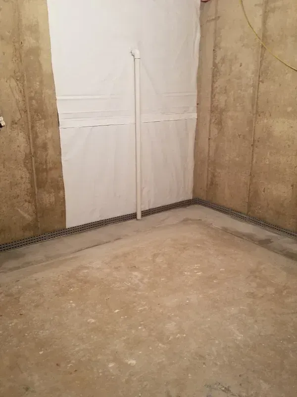 A basement corner featuring concrete walls, a white vapor barrier, and an exposed white PVC pipe running vertically.