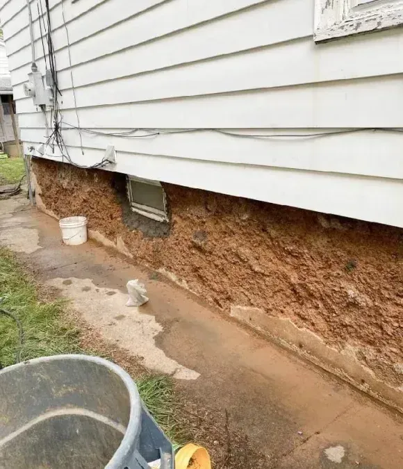 A side view of a white-sided house with a rough, exposed dirt and concrete foundation above a wet concrete walkway.