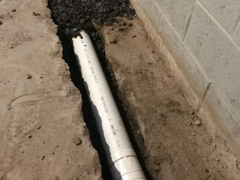 A white PVC pipe lies in a narrow, freshly dug trench alongside a concrete foundation wall filled with gravel.