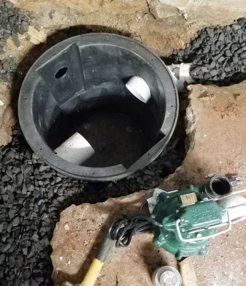 An open, black plastic sump pump basin set in gravel with PVC pipes connected, beside a green submersible sump pump.