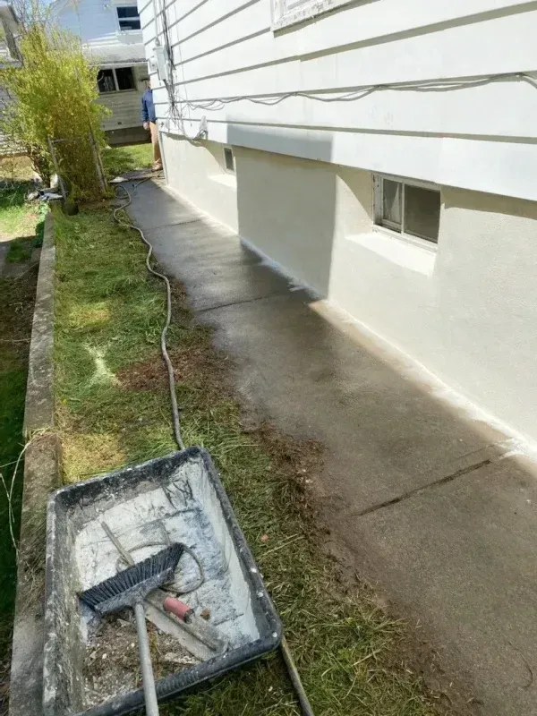 A white house foundation with a newly coated concrete path and a paint tray with a brush in the grass.