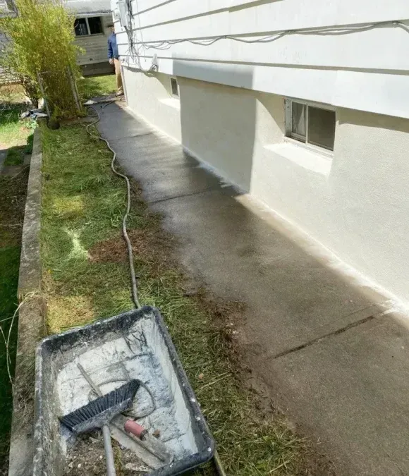 Freshly poured concrete walkway alongside the white foundation of a house with a utility tub and broom in the foreground.
