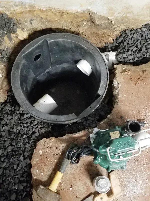 A black sump pump basin installed in a dirt hole with white PVC pipes, a green sump pump, and a hammer nearby.