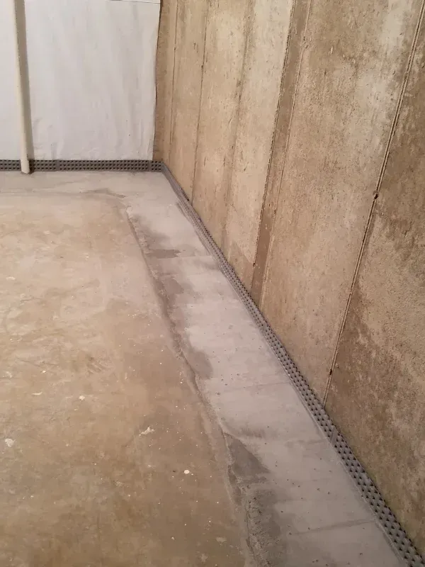 A concrete basement wall meeting a floor with a drainage channel installed along the corner perimeter.