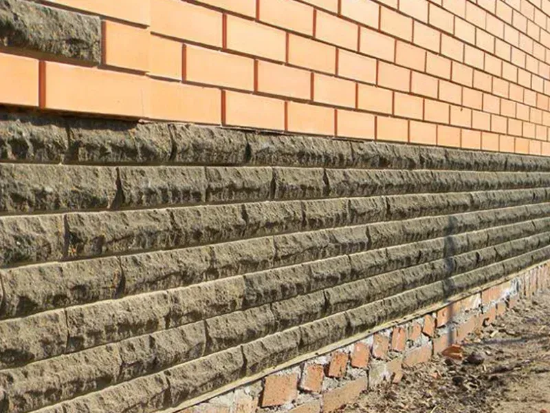 A low-angle view showing a tan brick wall sitting atop a foundation of horizontally stacked, textured dark stone blocks.