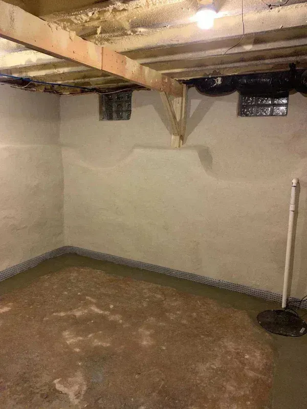 Unfinished basement corner with light-colored concrete walls, a wooden ceiling beam, and a dirt floor.