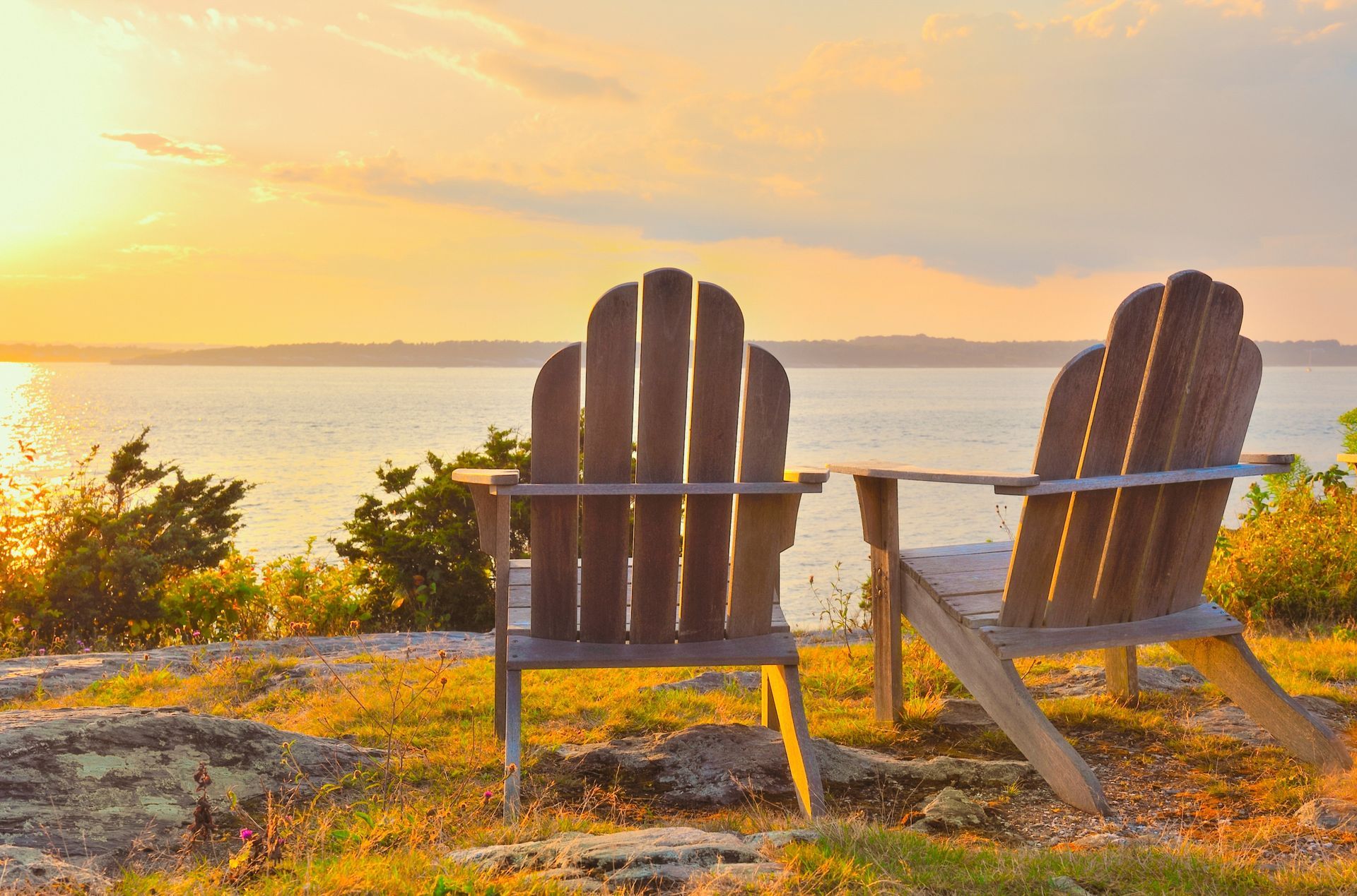 Two Adirondack chairs overlooking a calm lake at sunset, bathed in warm golden light.