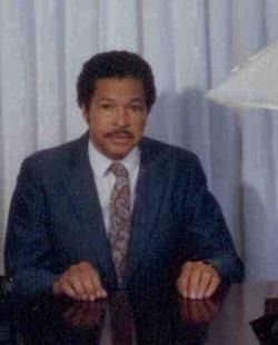 Man seated at a desk in a dark suit and tie, with curtains behind him.