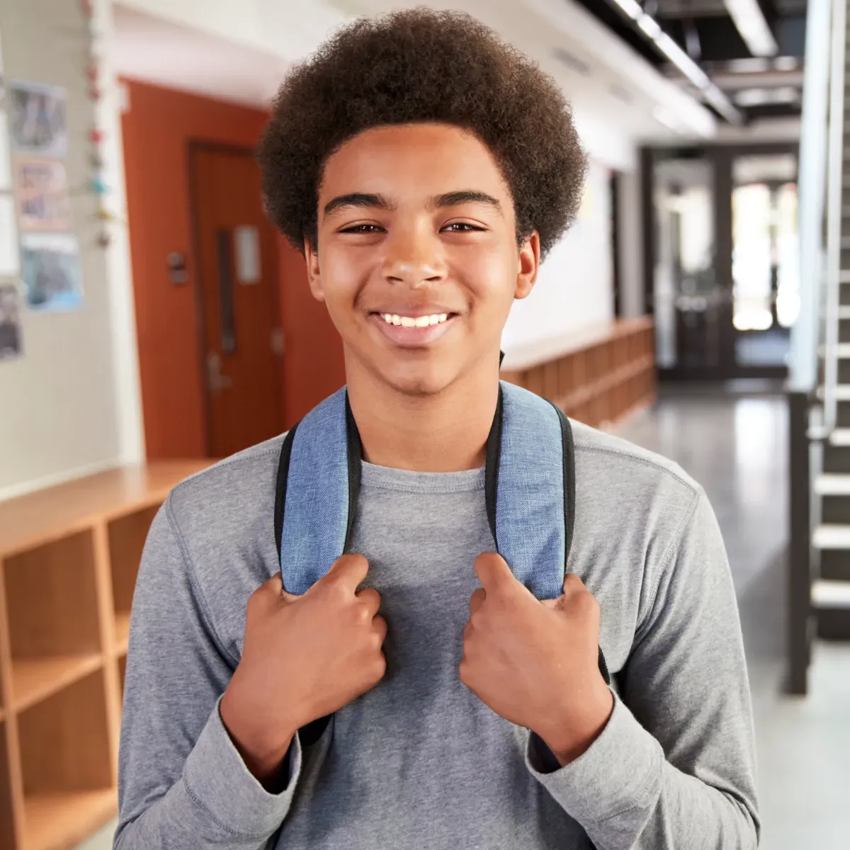 A young man wearing a backpack is smiling in a hallway.