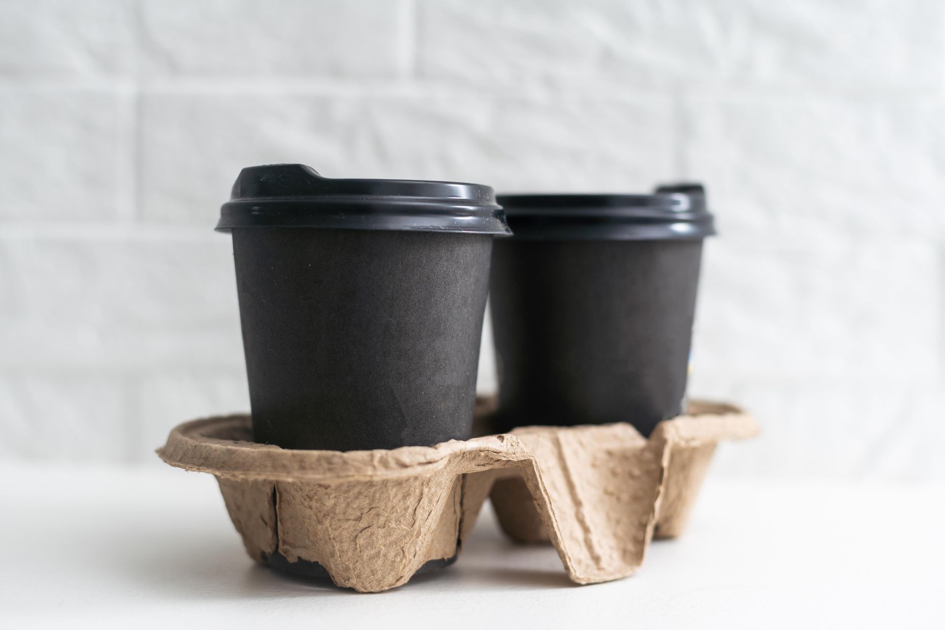 Two black coffee cups with lids in a cardboard carrier, against a white brick wall.
