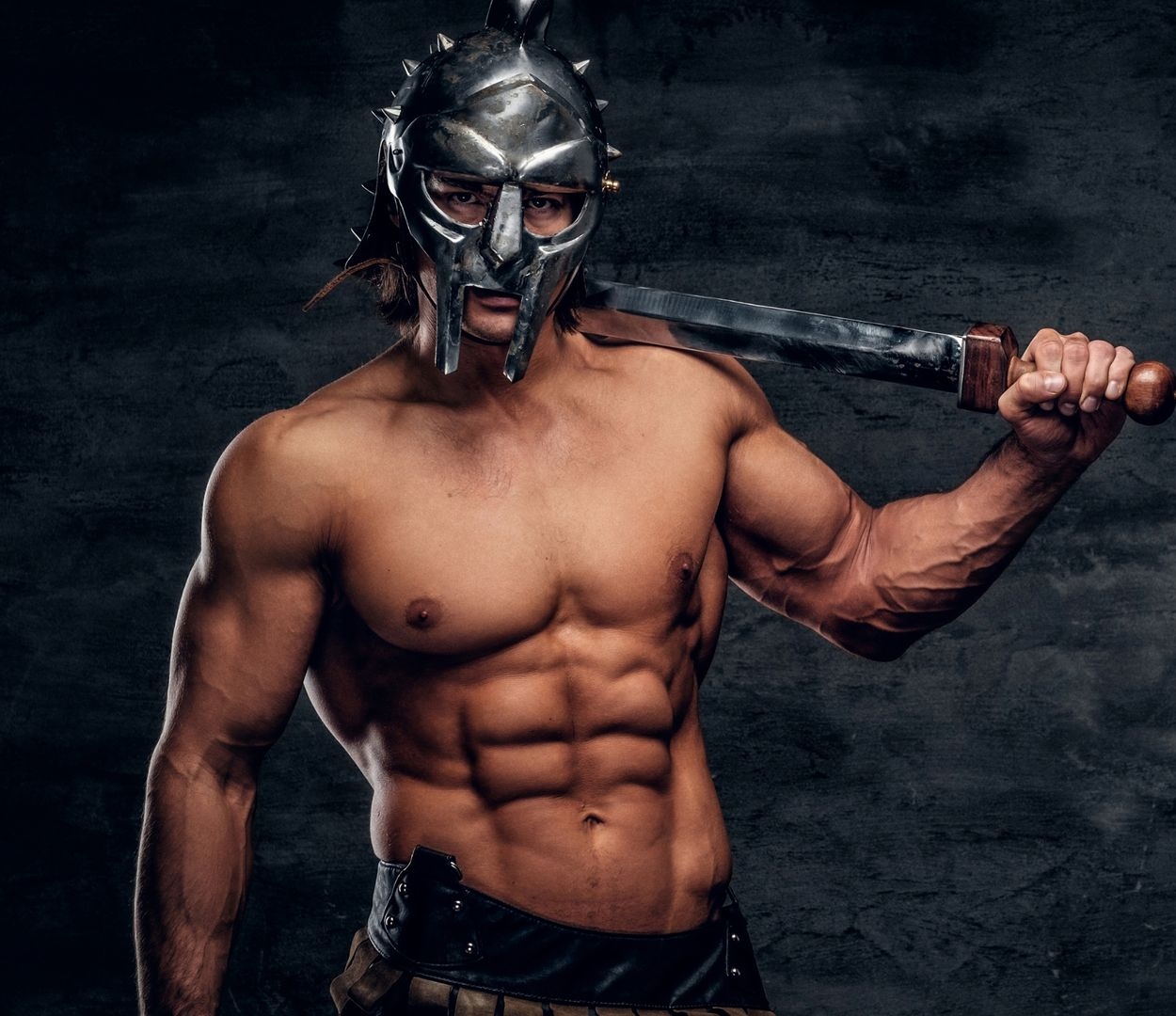 A muscular individual wearing a metal gladiator helmet, holding a sword over their shoulder against a dark background.