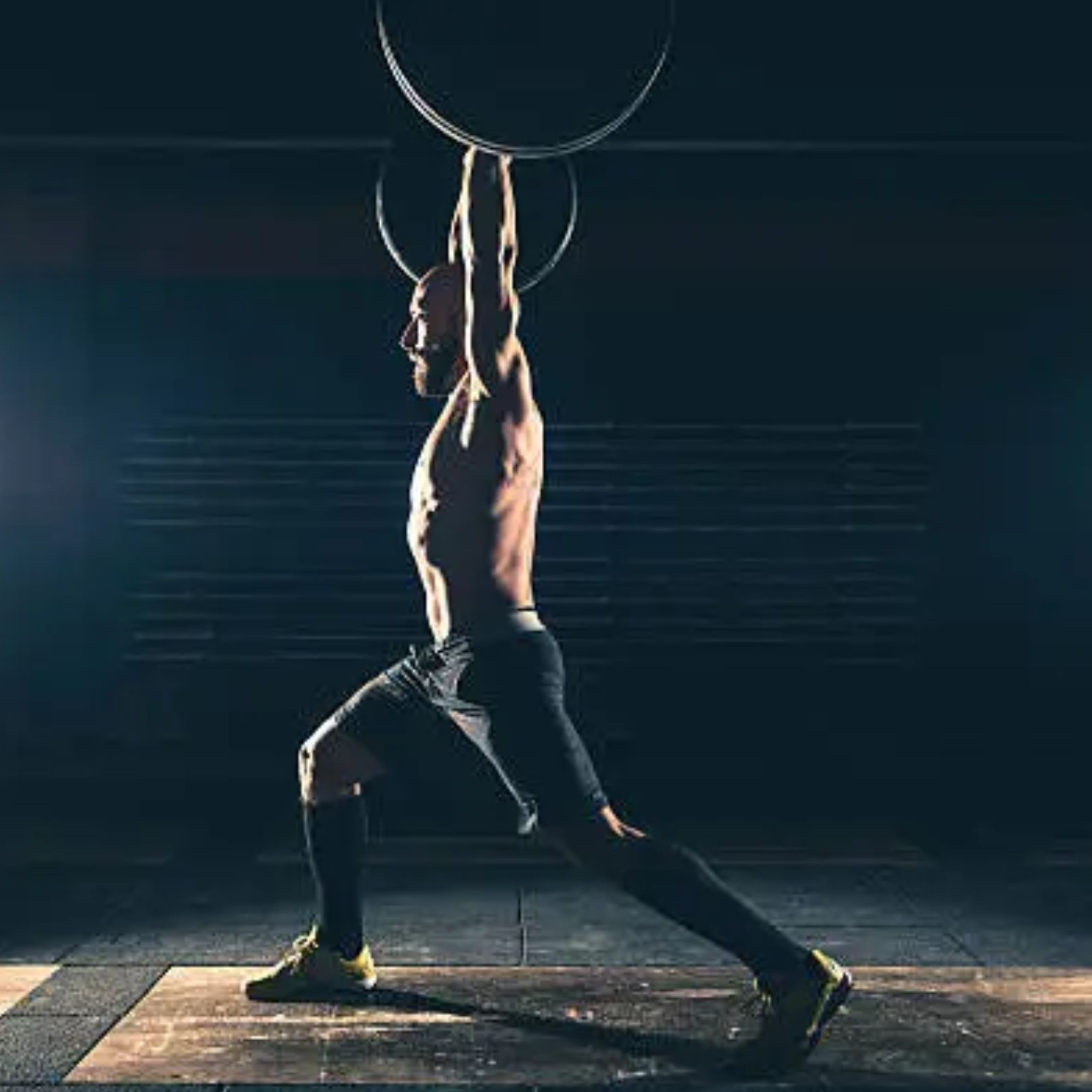 A shirtless athlete in a gym performing a barbell lunge with the weight held overhead.