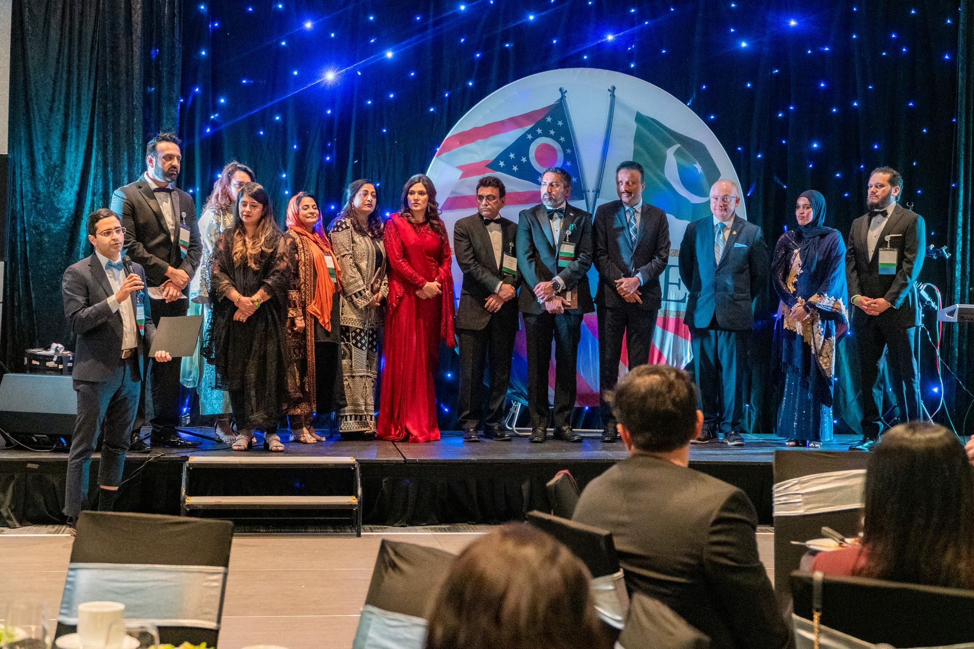 A large group of people in formal attire posing for a photo on a stage in front of a circular decorative backdrop.