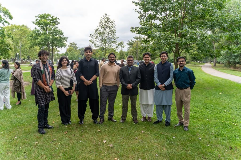 A group of eight people in formal attire standing in a row on a stage in front of a circular US and Pakistan flag backdrop.