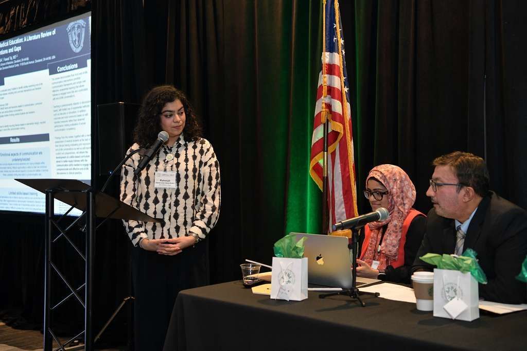 A group of eight people in formal attire standing in a row on a stage in front of a circular US and Pakistan flag backdrop.