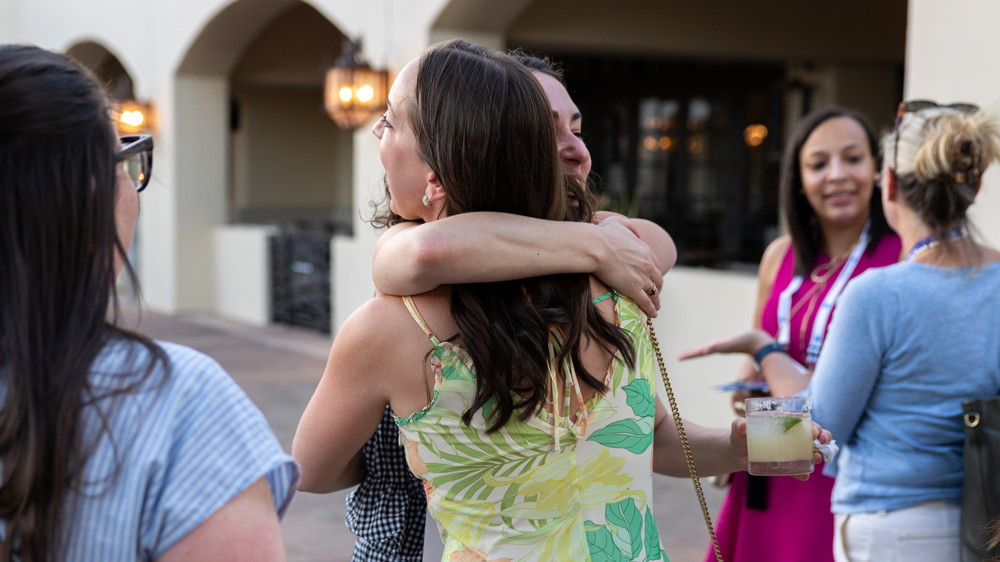 Two people hugging outdoors near a building, with others chatting nearby.