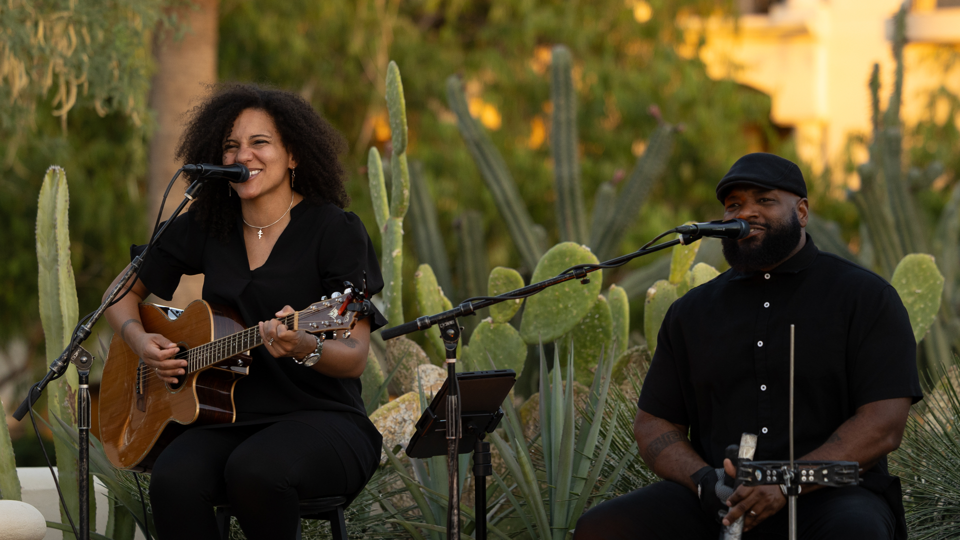 Two musicians performing outdoors, one singing with an acoustic guitar and the other at a mic.