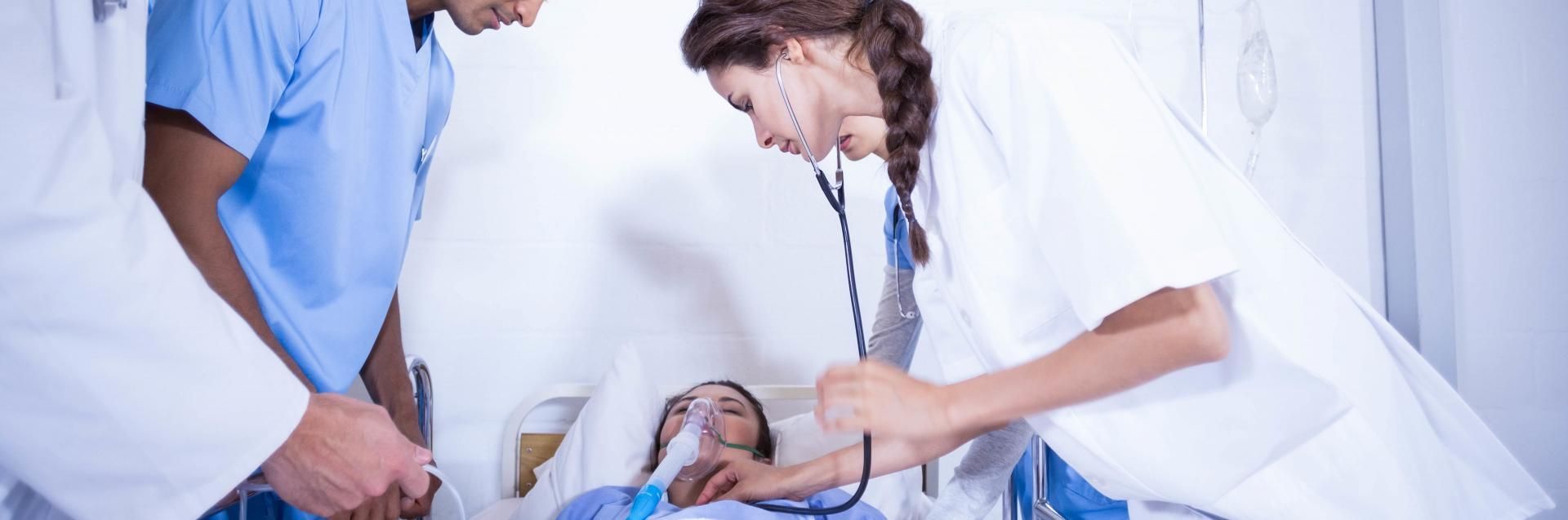 Doctor examining a patient with a stethoscope while another clinician stands nearby in a hospital room