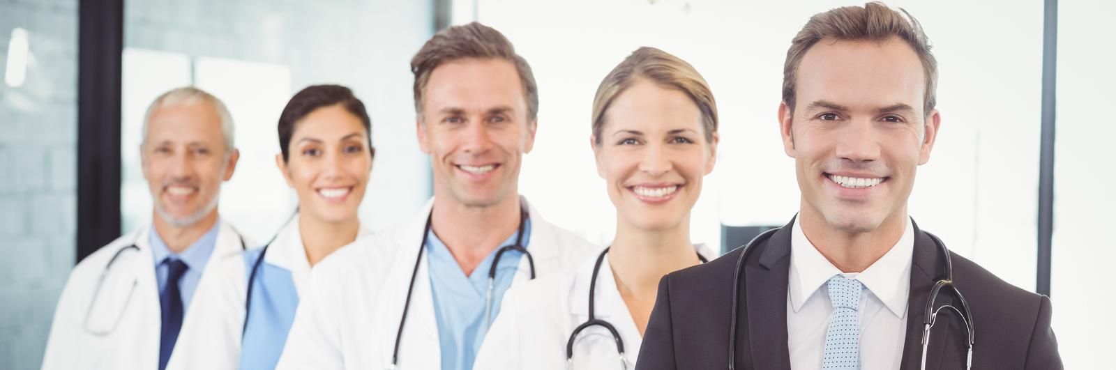 Six smiling medical professionals in white coats and scrubs standing in a bright office