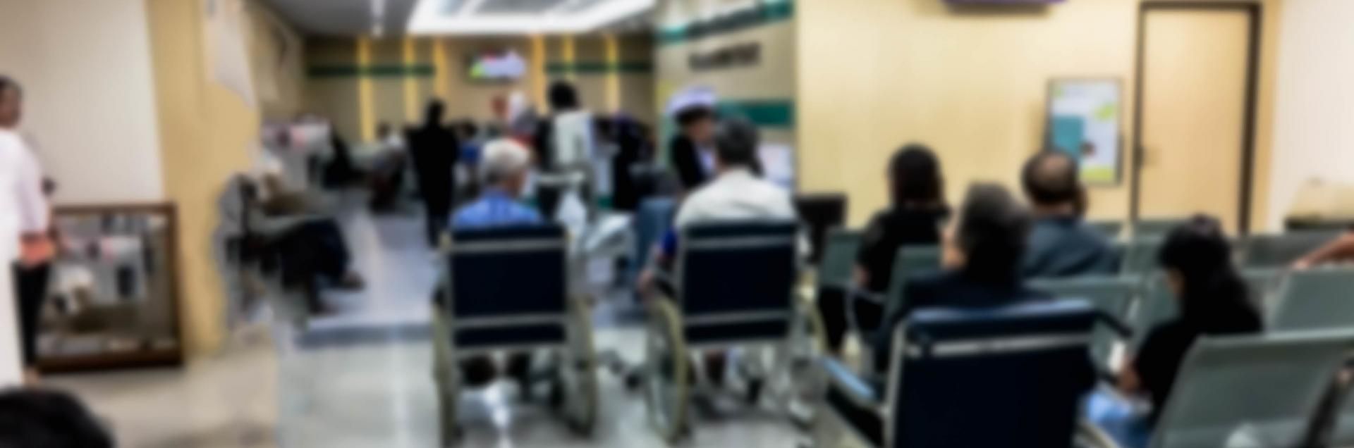 Busy waiting room with rows of chairs, people seated and standing, and a TV on the wall