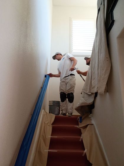 Two workers painting a stairwell, one on a ladder with blue tape along the wall.