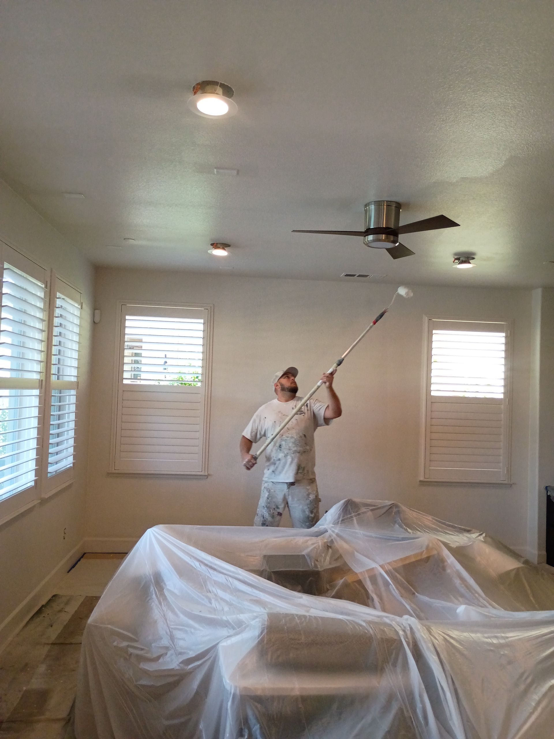Painter rolling white paint on a ceiling in a room covered with plastic sheeting.
