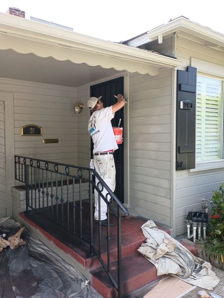 Person painting a front porch wall beside a doorway, with tools and supplies on the steps.