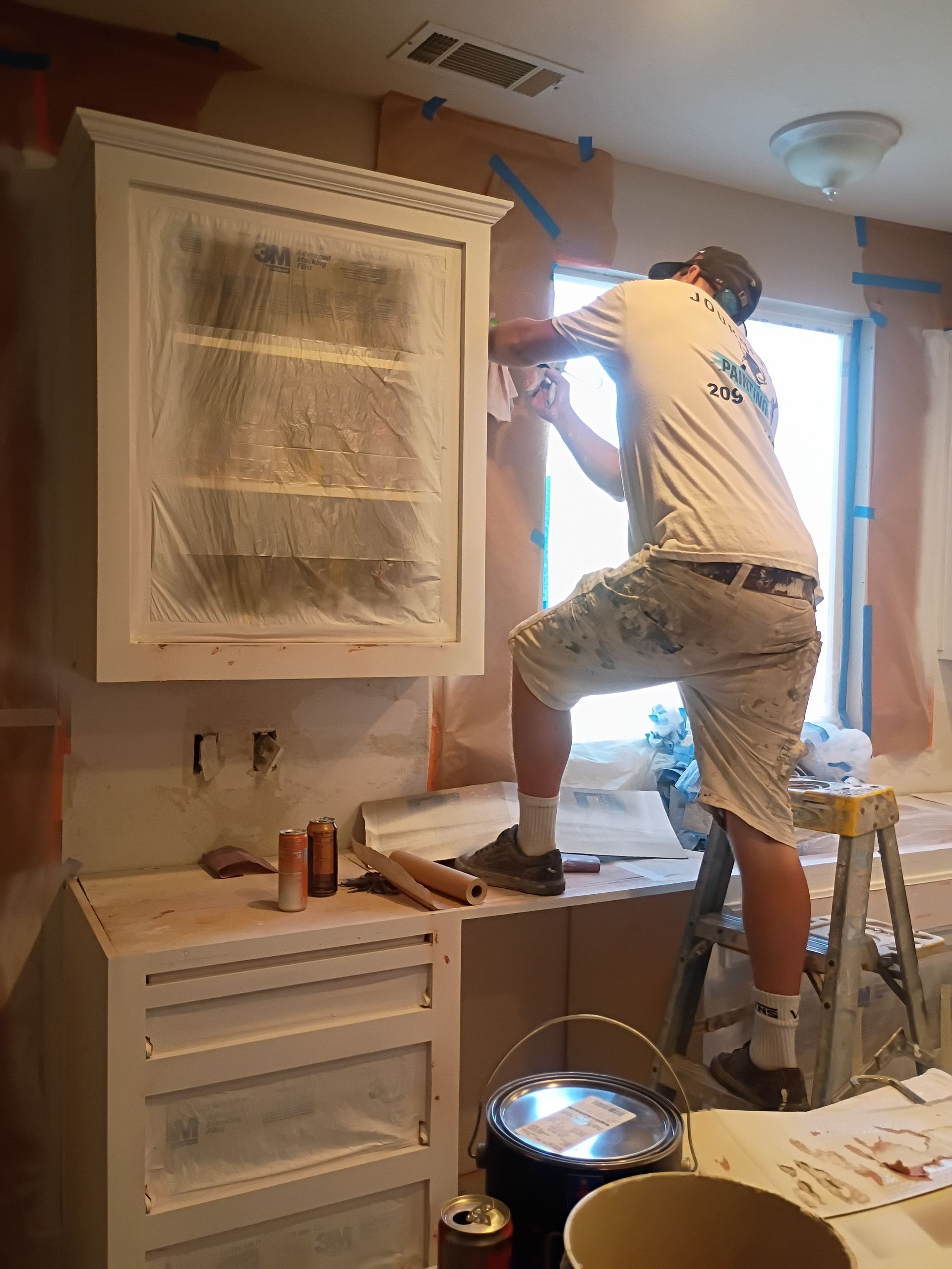 Person on a step stool painting a kitchen window frame and cabinets near a bright window.