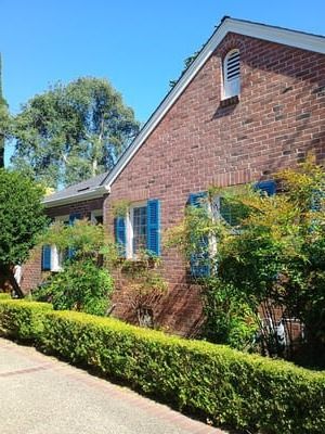 Brick house with blue shutters, white trim, and a neatly trimmed hedge along a sunny sidewalk.