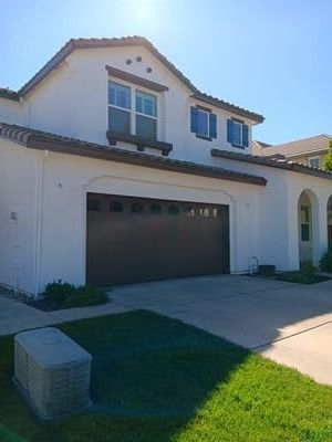 Suburban two-story house with stucco exterior, dark garage door, and front lawn in sunlight