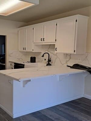 White kitchen with marble-look countertops, upper cabinets, black faucet, and dark wood flooring.