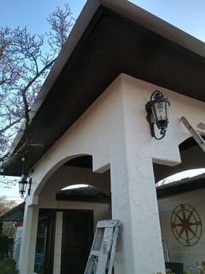 White stucco porch with dark roof, lantern lights, arched openings, and a ladder outside a house.