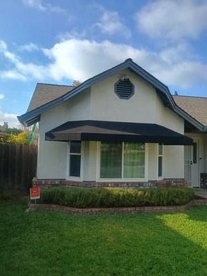 Small white house with gray roof, front porch awning, and green lawn under a blue sky.