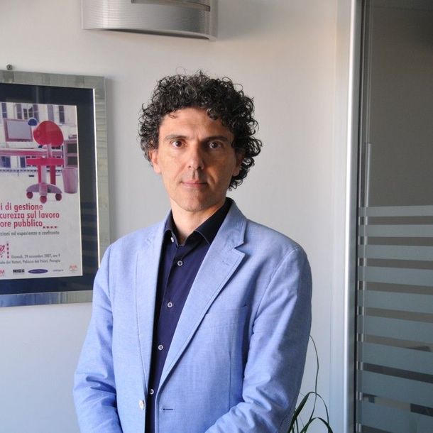 Uomo con blazer azzurro in piedi in un ambiente interno, accanto a una porta a vetri e a un poster appeso alla parete.
