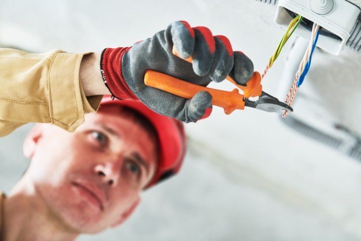Electrician in a red cap using pliers to strip insulation from wires hanging from a ceiling junction box.