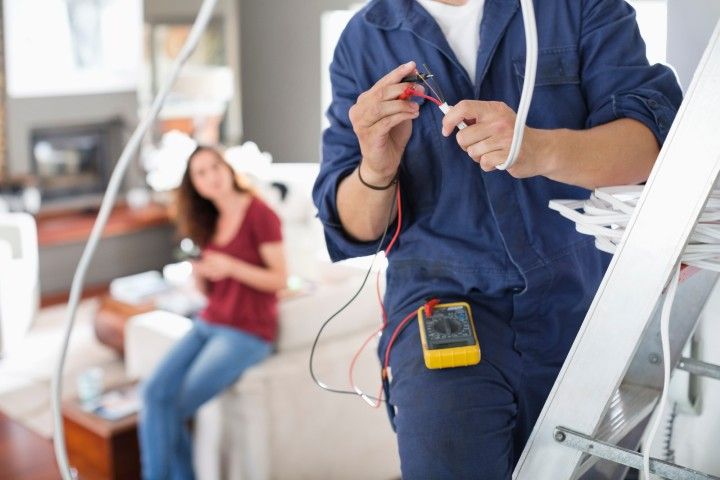 A technician wearing a blue uniform works with electrical wires and a multimeter while a person sits on a sofa nearby.