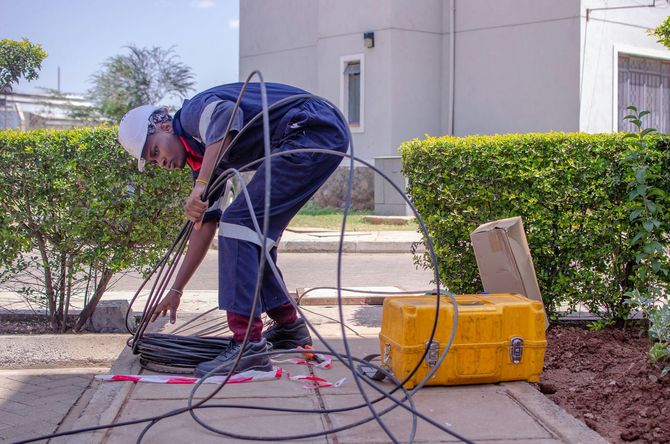 A worker in a hard hat and uniform inspects fiber optic cables at an outdoor ground access point near a yellow equipment box.