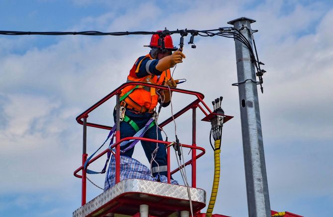 A utility worker in a high-visibility vest and hard hat works on power lines from an aerial lift bucket.