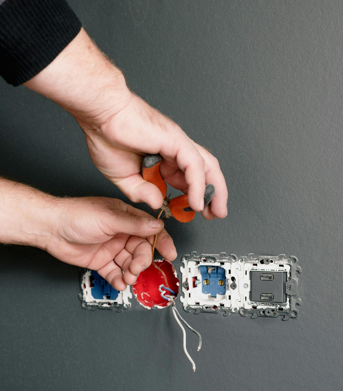 Hands using pliers to work with electrical wires at a wall-mounted outlet installation.