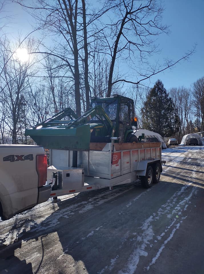 Un tracteur sur une remorque attelée à une camionnette, stationné dans une allée enneigée par une journée ensoleillée.