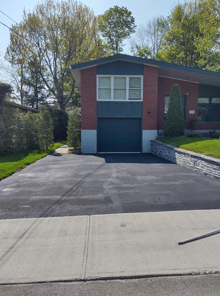 Allée asphaltée devant une maison en brique rouge avec garage noir.